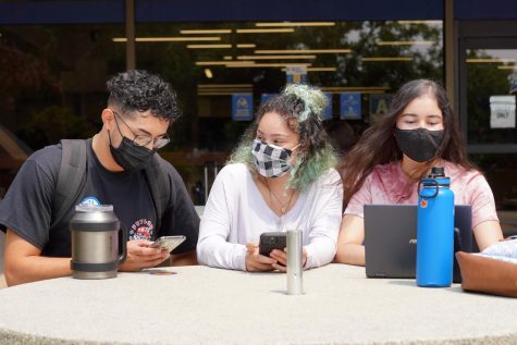 Three students with masks on interacting during the pandemic. All of them are wearing masks and one of them is using a computer.