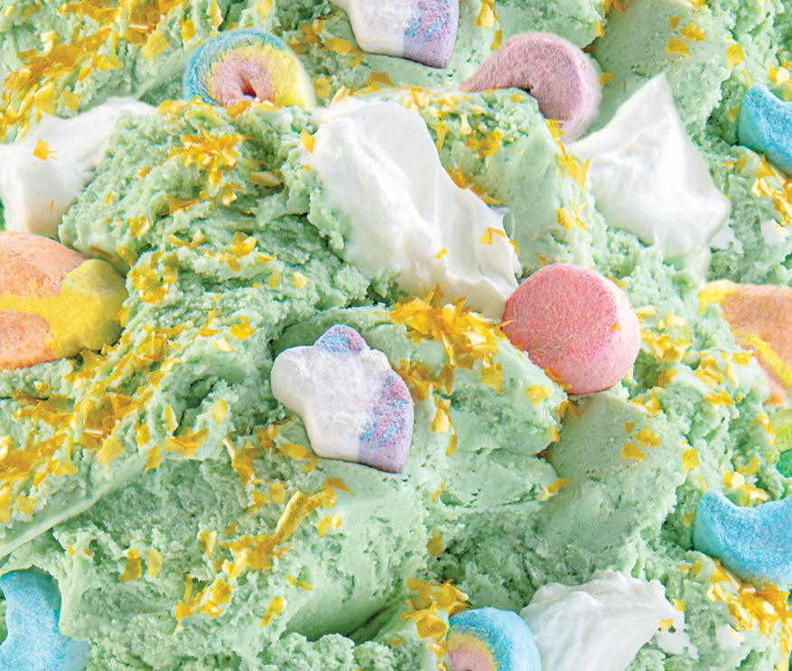 Cold Stone's "Ice Cream at the End of the Rainbow". A close-up of soft green ice cream mixed with pieces of Lucky Charm cereal, dollops of whipped topping, and gold glitter is shown. 
